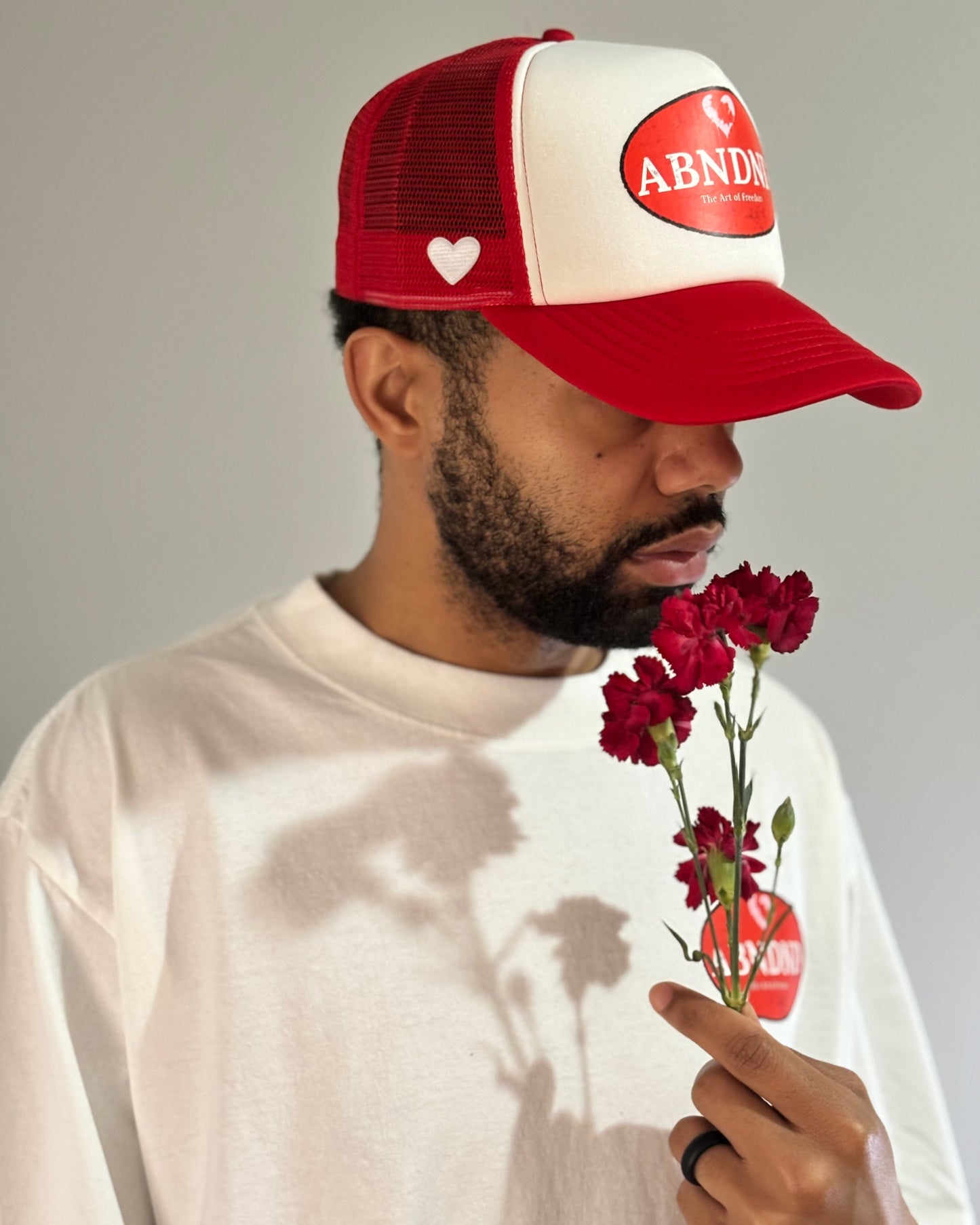 Man wearing a "Heart & Freedom" trucker hat with distressed ABNDND patch, mesh back, and holding red flowers.